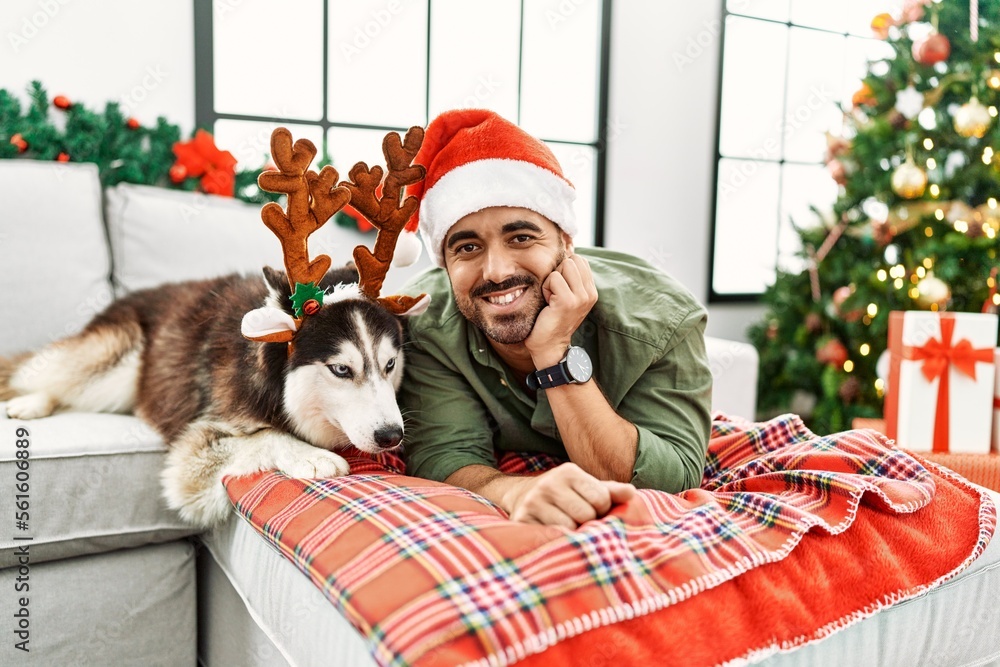 Obraz premium Young hispanic man lying on sofa with dog wearing reindeer hat by christmas tree at home