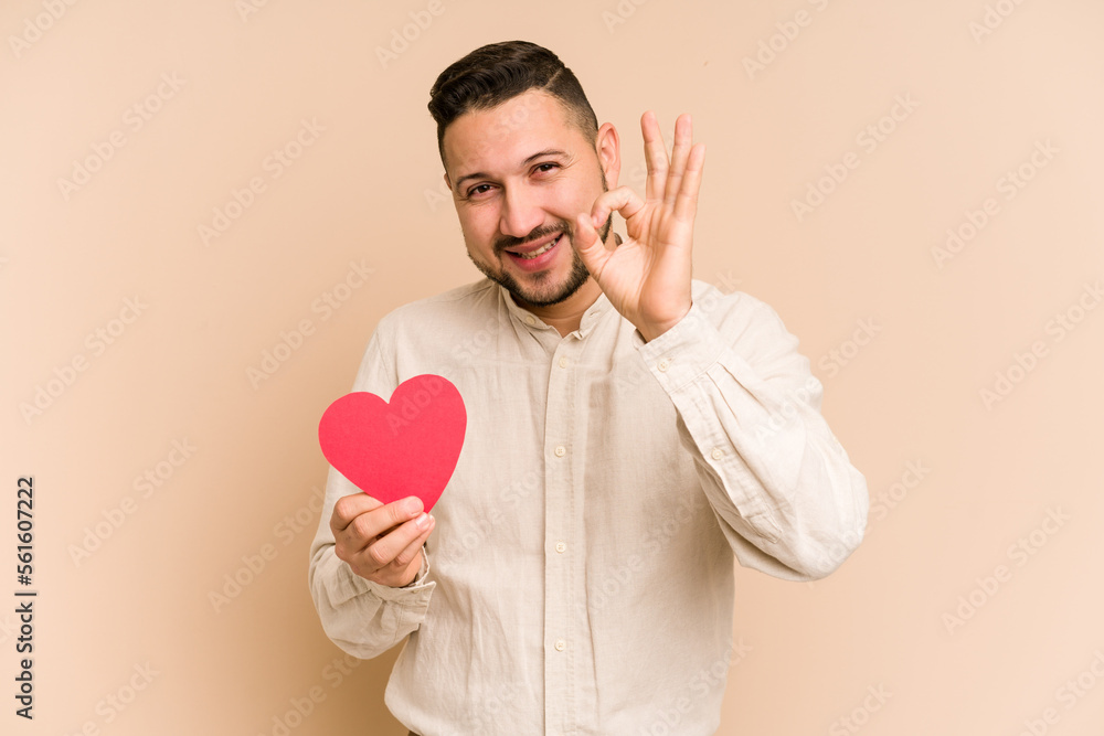 Adult latin man holding a red heart isolated cheerful and confident showing ok gesture. Adult latin man holding a red heart isolated cheerful and confident showing ok gesture.