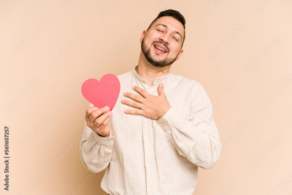 Adult latin man holding a red heart isolated laughs out loudly keeping hand on chest. Adult latin man holding a red heart isolated laughs out loudly keeping hand on chest.