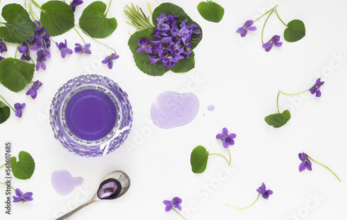 Syrup and jelly from viola odorata flowers in glass, spoon and violet fresh flower with leaves decorative arranged on white table background, shot from above. Spring herbal sweet product. Top view.