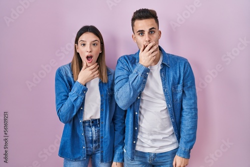 Young hispanic couple standing over pink background looking fascinated with disbelief, surprise and amazed expression with hands on chin