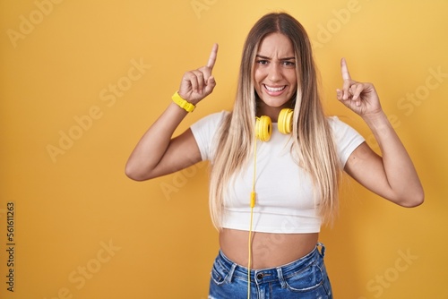 Young blonde woman standing over yellow background wearing headphones smiling amazed and surprised and pointing up with fingers and raised arms.