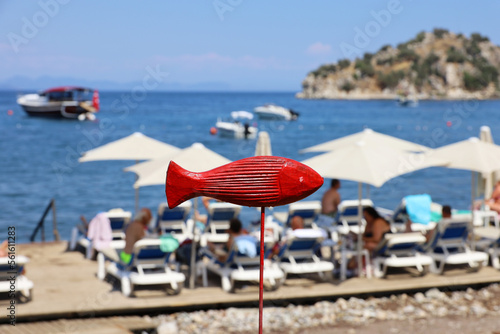 Selective focus to red wooden fish against sea beach with wicker parasols and sunbathing people. Mediterranean resort, motor boats and mountain in distance