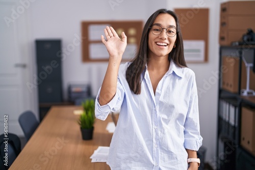 Young hispanic woman at the office waiving saying hello happy and smiling, friendly welcome gesture