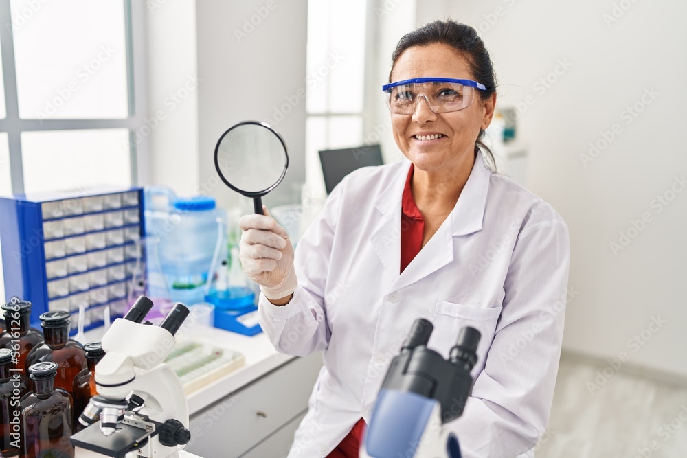 Middle age hispanic woman working at scientist laboratory holding magnifying glass looking positive and happy standing and smiling with a confident smile showing teeth