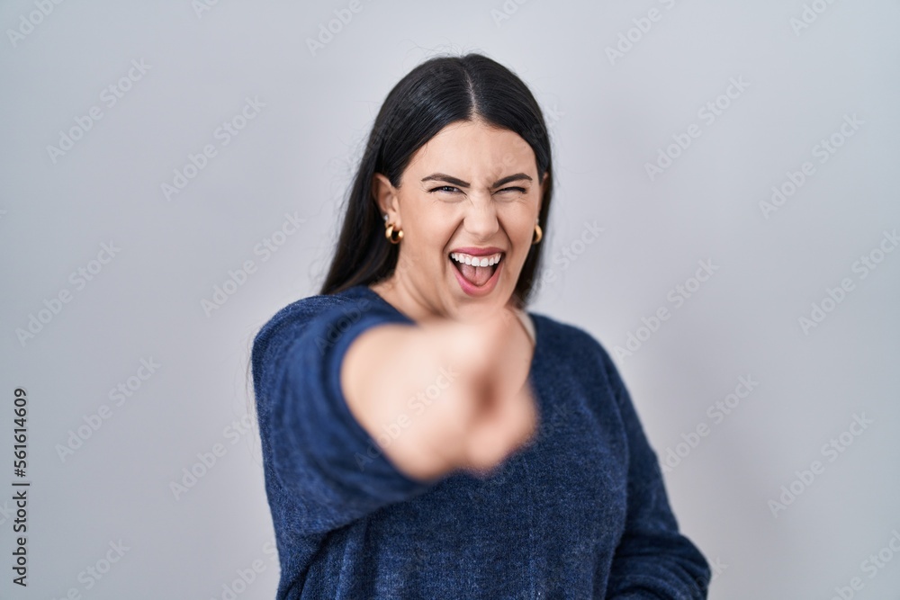 Young brunette woman standing over isolated background laughing at you, pointing finger to the camera with hand over body, shame expression Young brunette woman standing over isolated background laughing at you, pointing finger to the camera with hand over body, shame expression