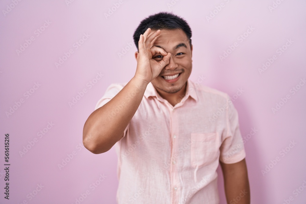 Chinese young man standing over pink background doing ok gesture with hand smiling, eye looking through fingers with happy face. Chinese young man standing over pink background doing ok gesture with hand smiling, eye looking through fingers with happy face.