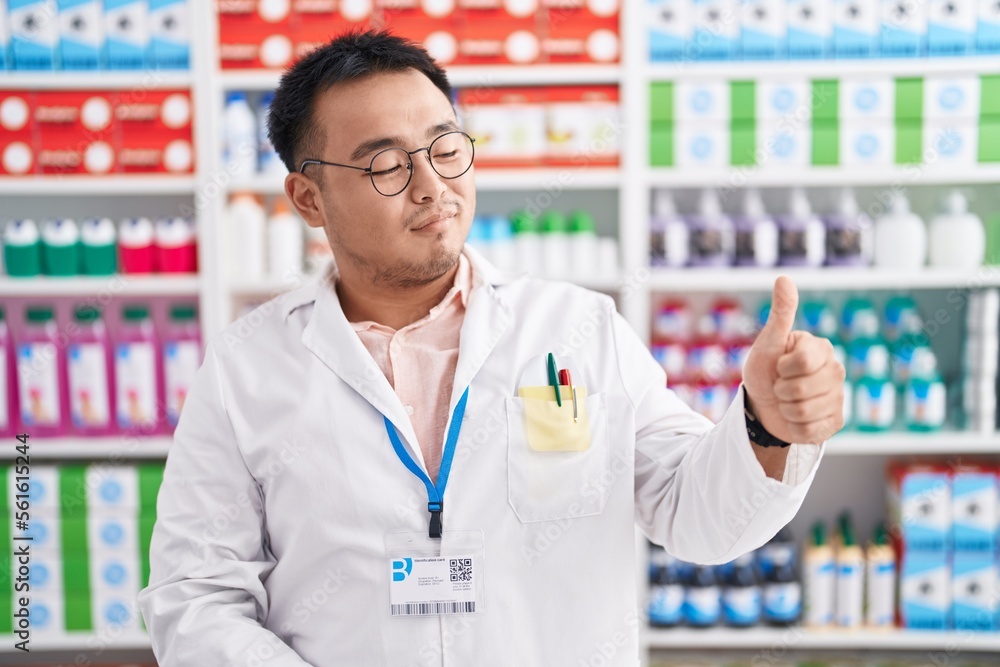 Chinese young man working at pharmacy drugstore looking proud, smiling doing thumbs up gesture to the side