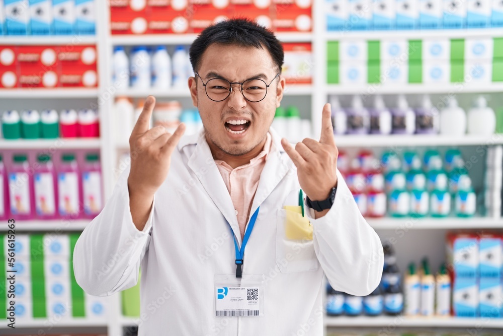 Chinese young man working at pharmacy drugstore shouting with crazy expression doing rock symbol with hands up. music star. heavy music concept. Chinese young man working at pharmacy drugstore shouting with crazy expression doing rock symbol with hands up. music star. heavy music concept.