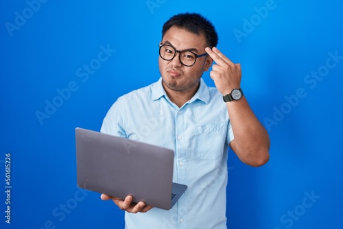 Chinese young man using computer laptop shooting and killing oneself pointing hand and fingers to head like gun, suicide gesture.