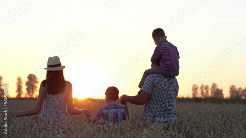 Parents silhouettes holding hands walk with lovely sons on wheat field at back sunset. Happy family explores countryside field and enjoys company