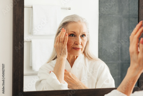 Aged woman looking at a mirror and touching her face. Female with grey hair wearing bathrobe standing in a bathroom.