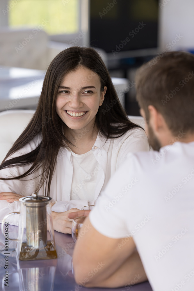 Obraz premium Positive beautiful young girl talking to boyfriend in cafe, looking at man, smiling, laughing. Couple dating over cup of tea, meeting for breakfast, enjoying love relationship, leisure, flirting