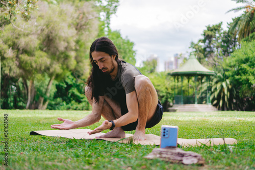 Concentrated young latin hippie man doing malasana pose on a yoga mat while filming himself in an open field with a gazebo behind. photo