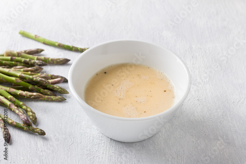 White ceramic bowl of beaten eggs with milk and fresh asparagus shoots on a light gray background. Cooking a delicious homemade omelet