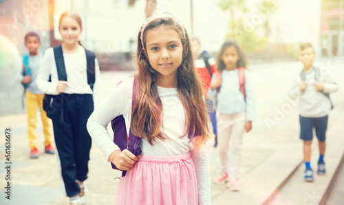 Portrait of positive schoolgirl standing near school, children on background