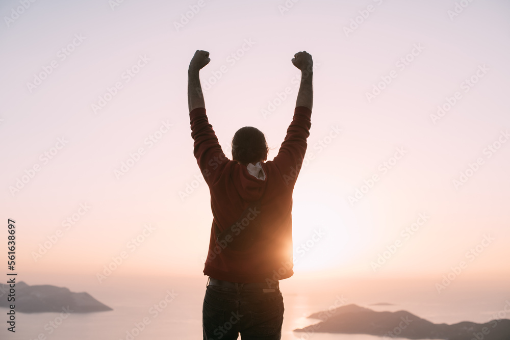 A young man meets the sunset, raising his hands in a victorious gesture on a mountain by the sea.