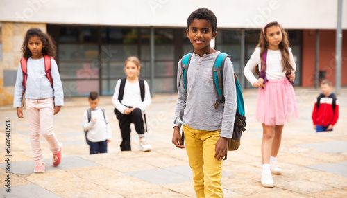 Cute tween african boy with backpack walking with other schoolchildren to school campus after lessons on warm fall day.