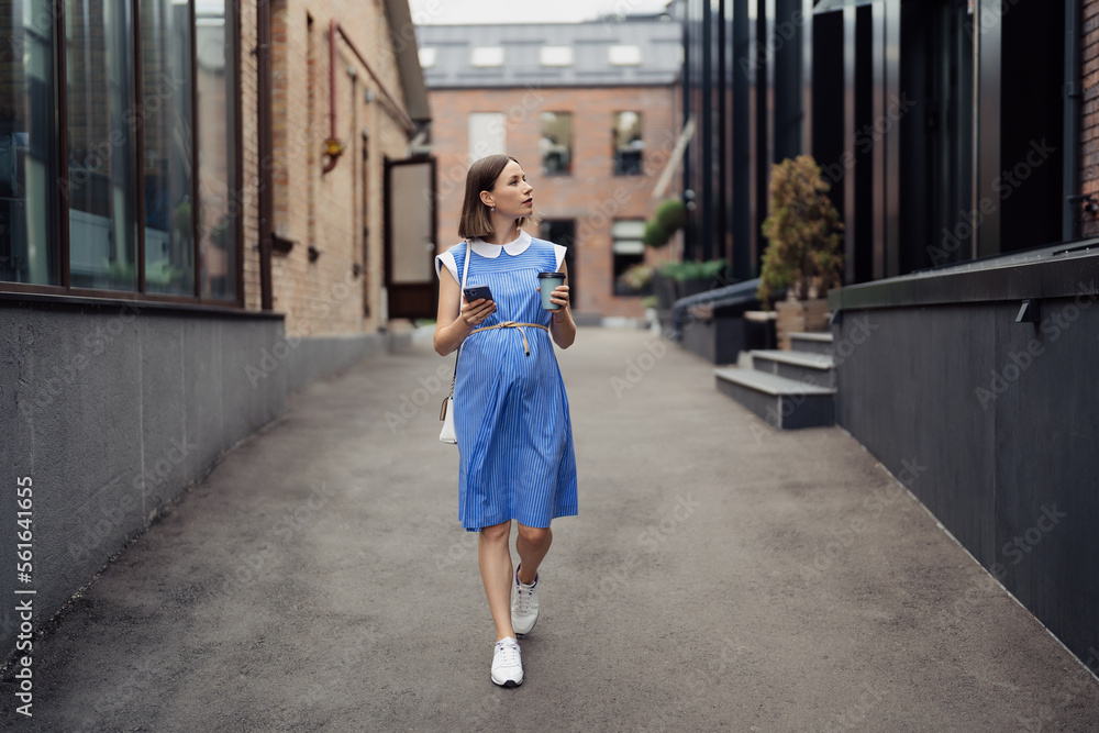 Busy modern pregnant woman walking among office buildings using a smartphone. Woman in a blue vintage dress and white purse using phone on the go