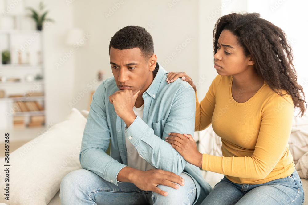 Apologize at home. Sad young african american female calms offended guy, support and caring, sit on sofa Apologize at home. Sad young african american female calms offended guy, support and caring, sit on sofa
