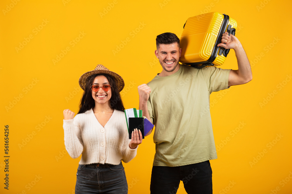 Smiling millennial arab woman in hat, glasses show passports, tickets, man with suitcase making victory hand gesture Smiling millennial arab woman in hat, glasses show passports, tickets, man with suitcase making victory hand gesture