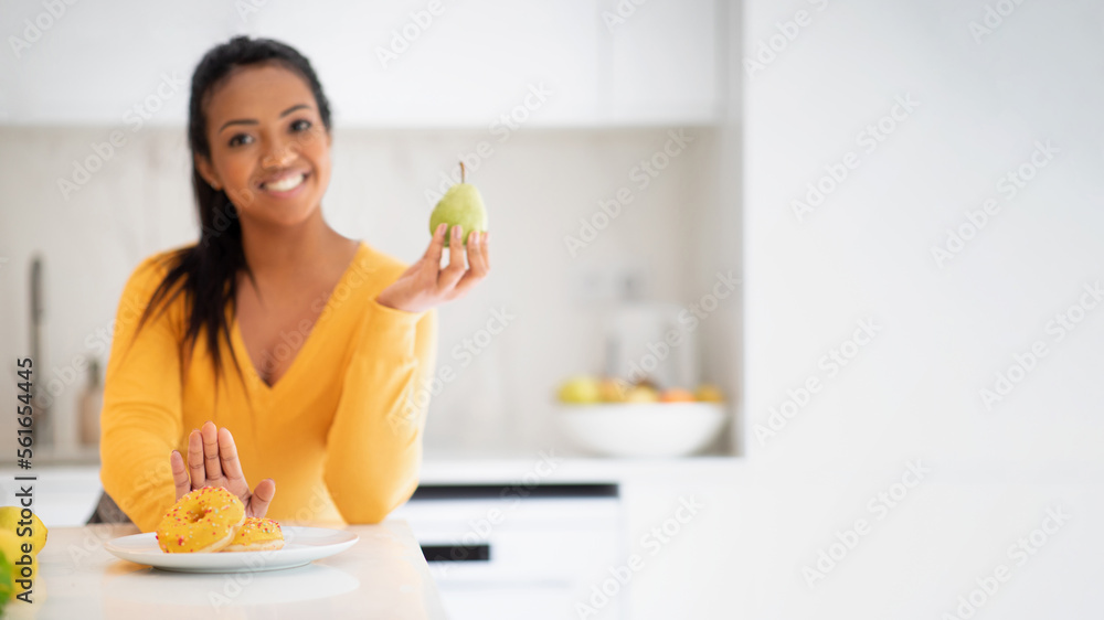 Cheerful young african american female recommending pear and refusing donut at table Cheerful young african american female recommending pear and refusing donut at table