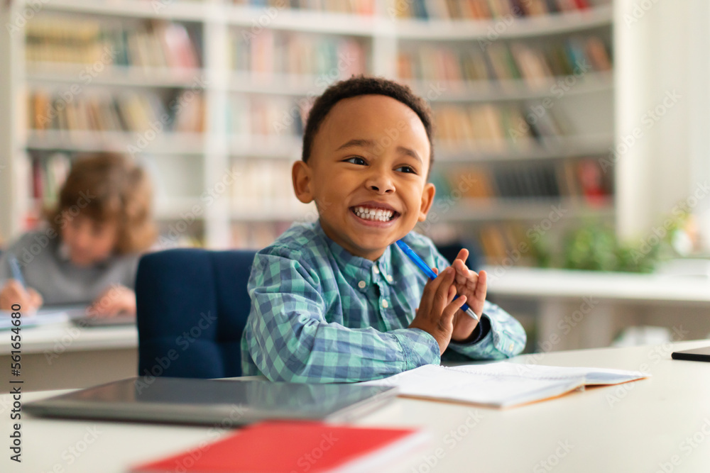 Happy african american primary school boy sitting at desk in classroom, writing in notebook and smiling, free space Happy african american primary school boy sitting at desk in classroom, writing in notebook and smiling, free space