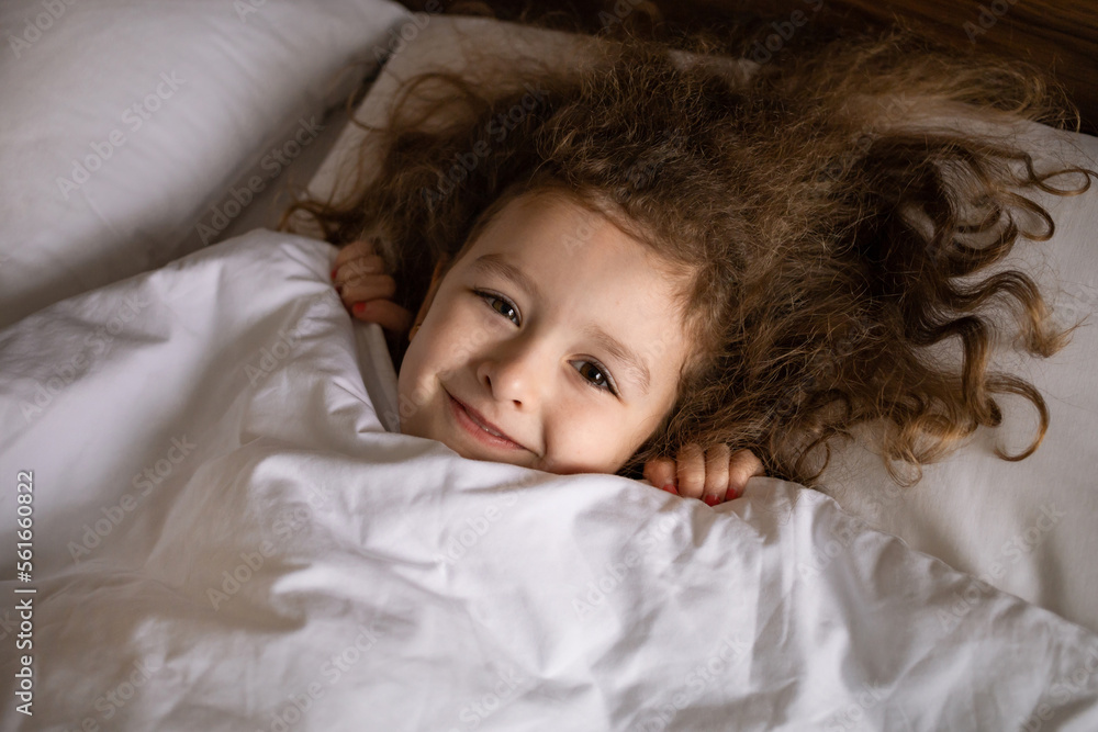 Happy child yawns, stretching her hands in bed. good morning baby. Adorable girl wakes up from sleep, looks out from under the blanket Happy child yawns, stretching her hands in bed. good morning baby. Adorable girl wakes up from sleep, looks out from under the blanket