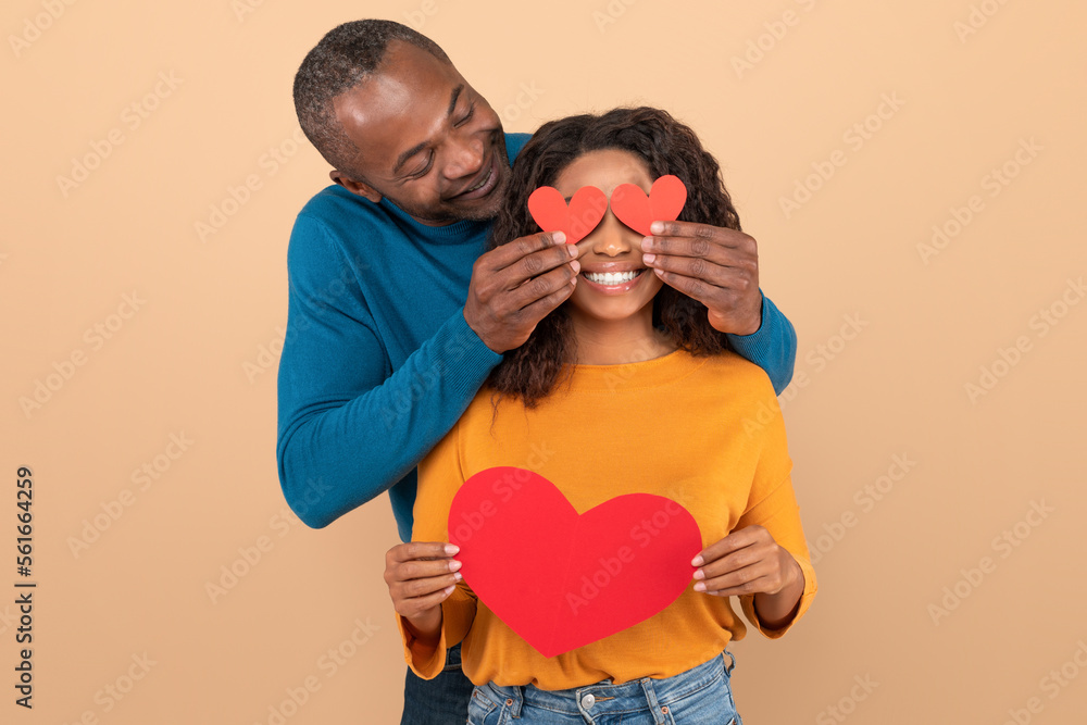Valentine's holiday celebration concept. Loving black middle aged man covering wife's eyes with paper hearts Valentine's holiday celebration concept. Loving black middle aged man covering wife's eyes with paper hearts