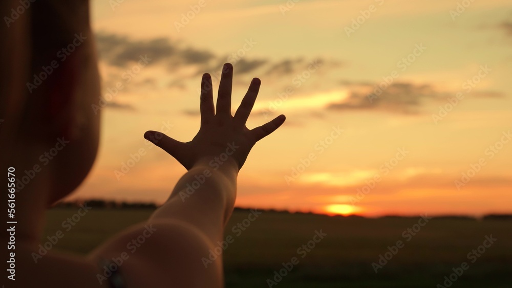 Hand of happy child girl reaches for sunset and beautiful sky. Sun between fingers of kids hand in park. Little girl stretches her hand to sun, dreams in nature. Child plays with sun. Happy family.