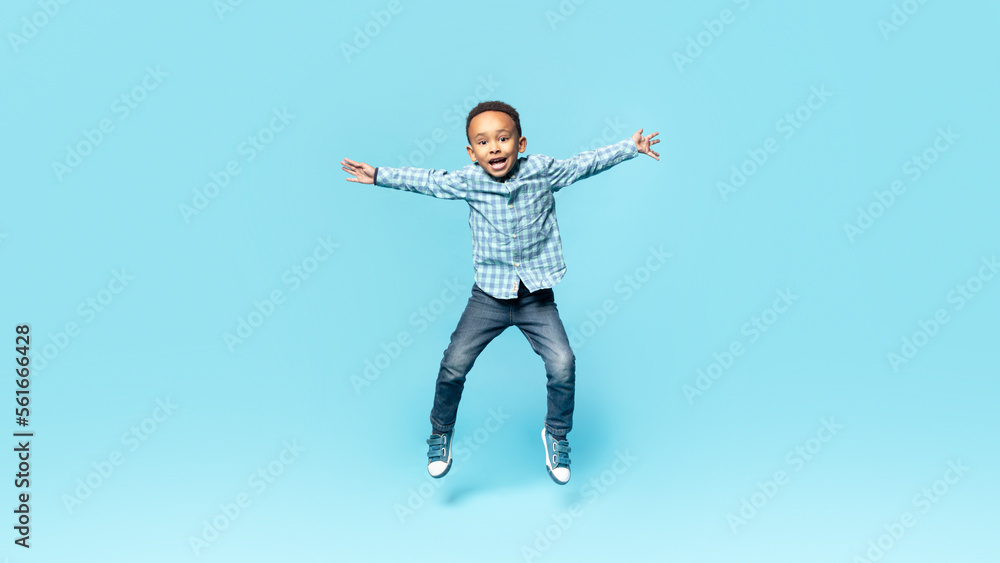 Happy little african american boy posing in mid air, jumping and spreading arms over blue studio background, full length