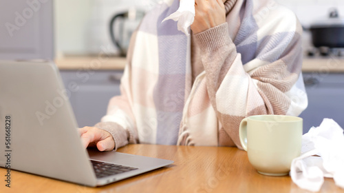 A woman wipes her face with a paper napkin and works at a laptop. A woman is sick and working remotely at home at the kitchen table. Napkins, runny nose, flu, orvi, sick leave, cold, photo