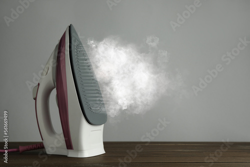 Modern iron with steam on wooden table against light grey background, space for text