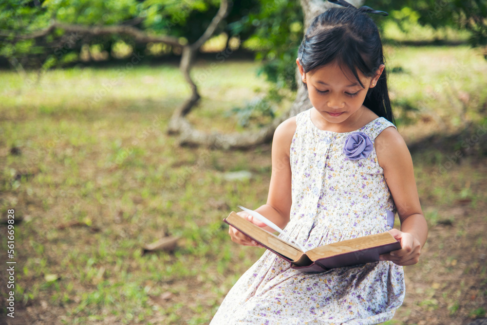 Asian Girl holding book reading at green park in natural garden. Young todler girl relaxation read open book self study. Happy child women smiling with happiness learning. Kid sit in green park