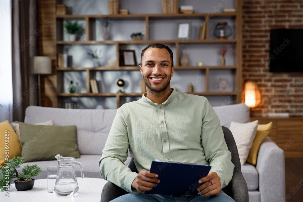 Young African American man sitting at home in a stylish office. Psychologist, coach or businessman looking at the camera and smiling. Beautiful teeth and smile. Young African American man sitting at home in a stylish office. Psychologist, coach or businessman looking at the camera and smiling. Beautiful teeth and smile.