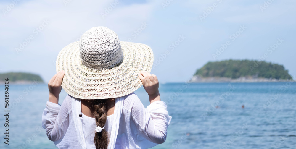 Fototapeta premium A Caucasian girl in a straw hat and a white tunic stands with her back to the camera and looks at the sea. Space for text.