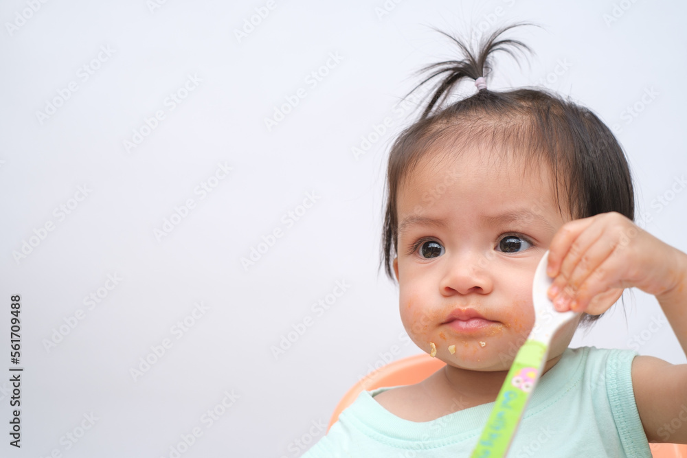 Cheerful baby child sitting and eats food by herself with spoon. Portrait of happy kid boy or girl in high chair. Smiling feeding delicious food concept. Isolate on white background. Cheerful baby child sitting and eats food by herself with spoon. Portrait of happy kid boy or girl in high chair. Smiling feeding delicious food concept. Isolate on white background.