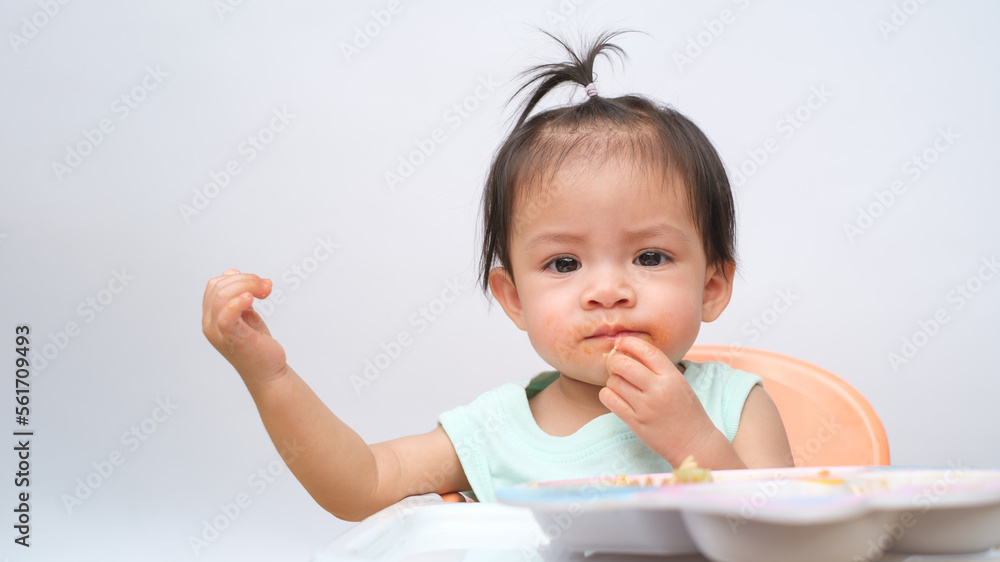 Cheerful baby child sitting and eats food by herself with spoon. Portrait of happy kid boy or girl in high chair. Smiling feeding delicious food concept. Isolate on white background. Cheerful baby child sitting and eats food by herself with spoon. Portrait of happy kid boy or girl in high chair. Smiling feeding delicious food concept. Isolate on white background.