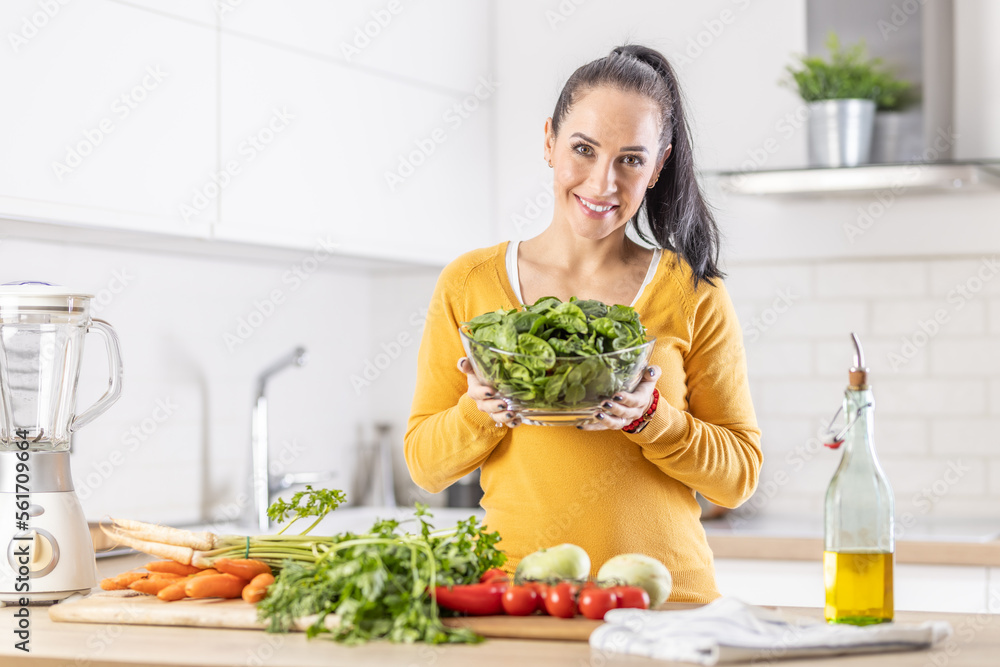 Smiling woman with a bowl full of spinach leaves along with fresh vegetables and olive oil in her kitchen