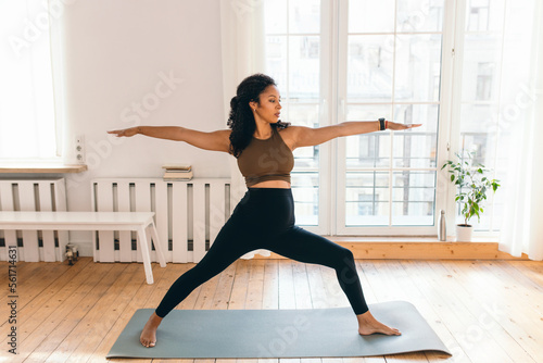 Pregnant woman doing prenatal yoga at home, standing in warrior posture on mat in her living-room against big window, outstretching hands, wearing smart watch to control pulse, heart rate and calories