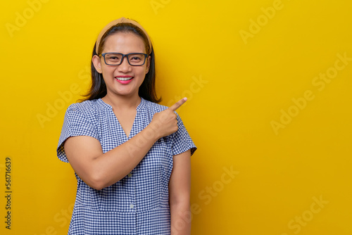 Fototapeta Naklejka Na Ścianę i Meble -  cheerful young Asian woman wearing a dress checkered with glasses pointing a finger at an empty room space isolated on yellow background. people lifestyle concept