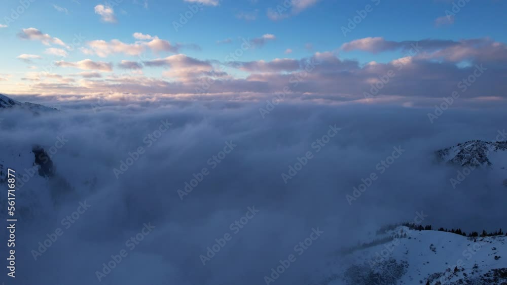 Flying among the ocean of clouds in the mountains. The fog is like big ocean waves crashing against high mountains covered with snow. Spruce trees grow in places. Sunset. Orange rays of the sun Flying among the ocean of clouds in the mountains. The fog is like big ocean waves crashing against high mountains covered with snow. Spruce trees grow in places. Sunset. Orange rays of the sun