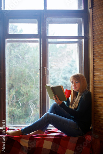 Charming young dreamy girl sits on a window with a book and cup.
