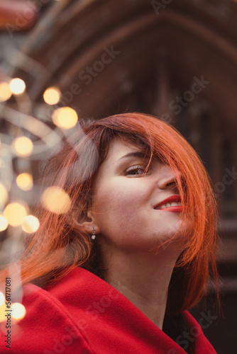 Close up attractive redhead lady and fairy lights on street portrait picture. Closeup side view photography with old building on background. High quality photo for ads, travel blog, magazine, article