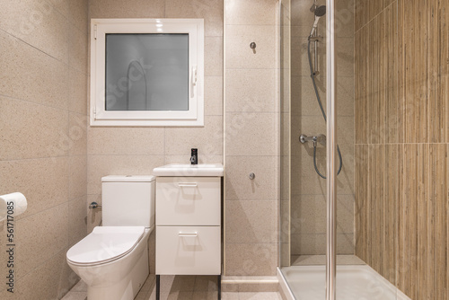 Interior of compact bathroom with beige marble tiles. Toilet next to the washbasin on small vanity and window in the wall for natural ventilation. Shower area is separated by glass railing.