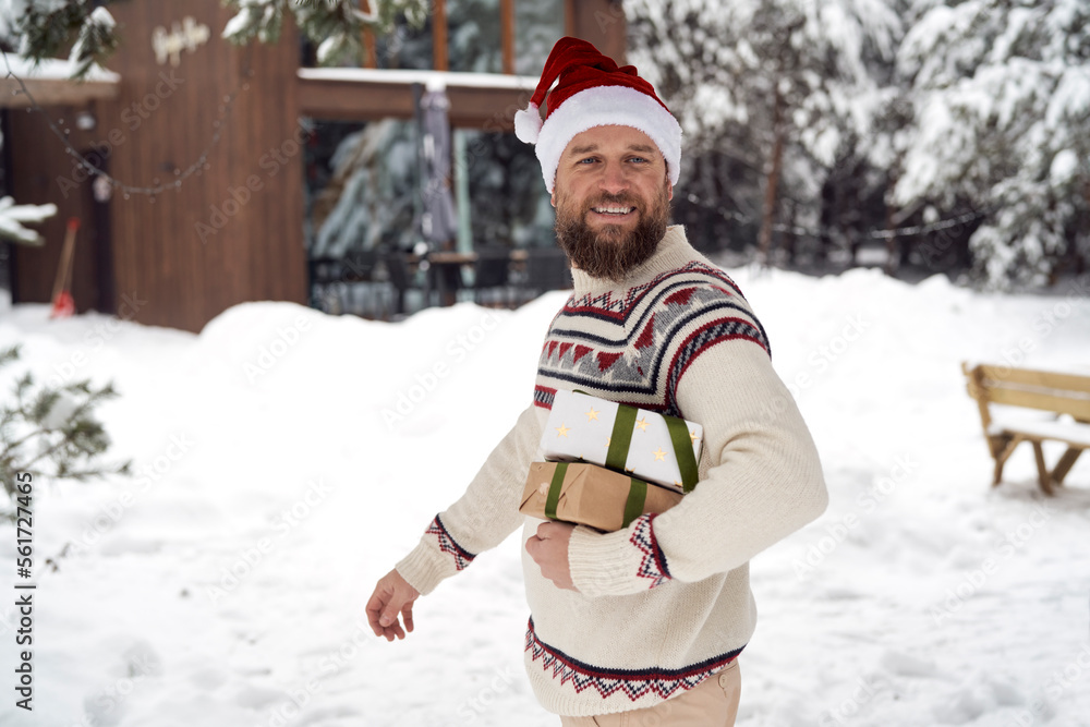 Fototapeta premium Caucasian man with Santa hat holding Christmas present in front of house in winter time