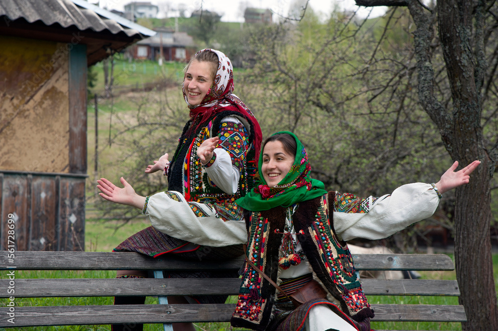 Young girls dressed in hutsul national clothes sitting on a bench and have fun talking in front of the hut. Ukraine.