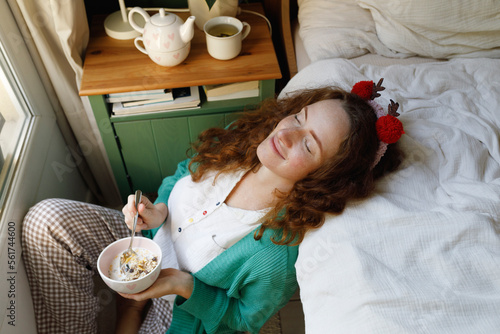 Smiling woman with eyes closed holding cereal bowl by bed at home photo