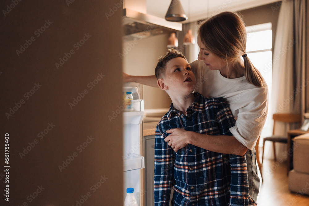 Young mother standing with her little son near fridge in kitchen Young mother standing with her little son near fridge in kitchen