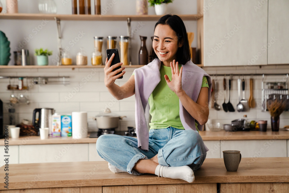 Smiling asian woman making video call via mobile phone and waving while sitting in kitchen Smiling asian woman making video call via mobile phone and waving while sitting in kitchen